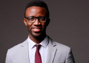 Professional headshot of a smiling man in a light gray suit, white shirt, burgundy tie, and glasses against a dark backdrop.