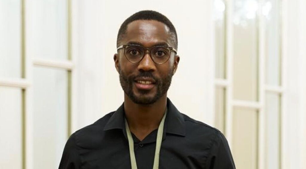 Felix Naanda - The Brief | Namibia's Leading Business & Financial News Portrait of a man with short hair and a beard wearing round glasses and a black button-down shirt in a bright hallway.