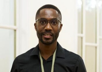 Portrait of a man with short hair and a beard wearing round glasses and a black button-down shirt in a bright hallway.