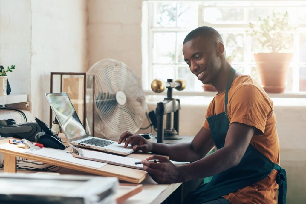 Smiling man in an orange shirt and teal apron sits at a wood-workbench, typing on a laptop amid papers and tools in a bright workshop.