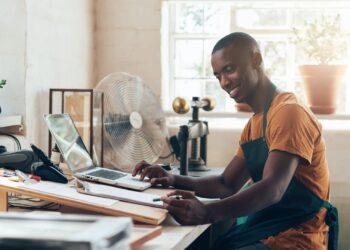 Smiling man in an orange shirt and teal apron sits at a wood-workbench, typing on a laptop amid papers and tools in a bright workshop.