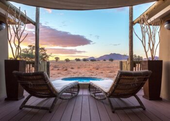 Two wicker chaise lounges on a wooden deck face a desert landscape with a small circular pool and a sunset sky over distant mountains.