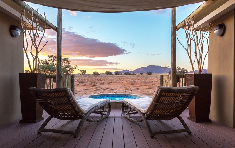 Two wicker chaise lounges on a wooden deck face a desert landscape with a small circular pool and a sunset sky over distant mountains.
