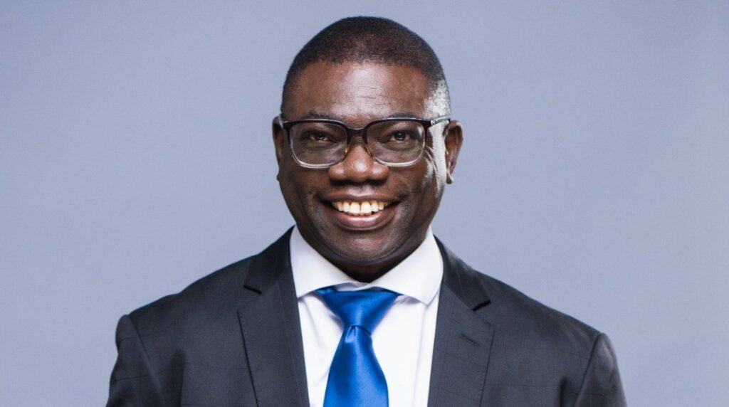 Evans Simataa - The Brief | Namibia's Leading Business & Financial News Smiling man in a dark suit and blue tie against a light gray backdrop, wearing glasses.