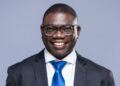 Evans Simataa - The Brief | Namibia's Leading Business & Financial News Smiling man in a dark suit and blue tie against a light gray backdrop, wearing glasses.