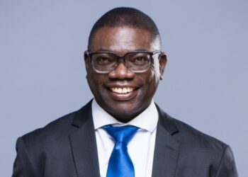 Smiling man in a dark suit and blue tie against a light gray backdrop, wearing glasses.