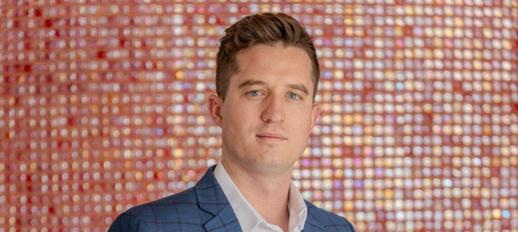 Portrait of a man wearing a blue checkered suit and white shirt, looking at the camera against a colorful bokeh background.
