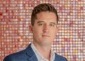 Portrait of a man wearing a blue checkered suit and white shirt, looking at the camera against a colorful bokeh background.