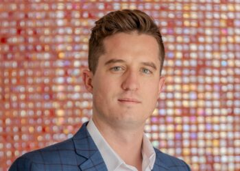 Portrait of a man wearing a blue checkered suit and white shirt, looking at the camera against a colorful bokeh background.