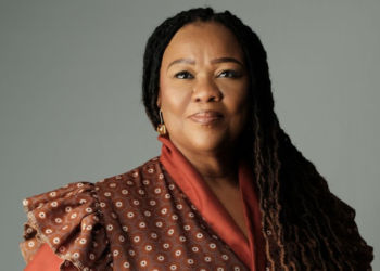 Portrait of a woman with long braids wearing a red patterned blouse against a gray background.