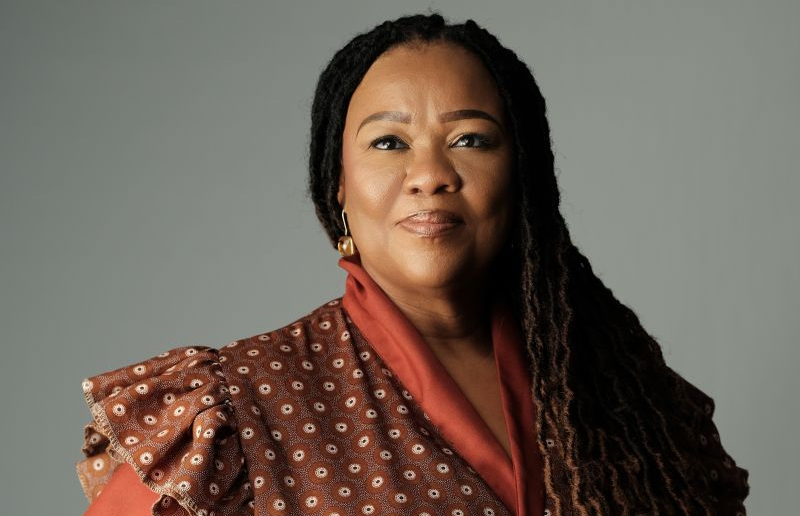 ZENZIE AWASES - The Brief | Namibia's Leading Business & Financial News Portrait of a woman with long braids wearing a red patterned blouse against a gray background.