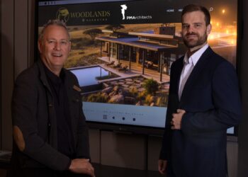 Two men stand beside a large screen showing a modern house rendering with Woodlands branding in a showroom setting.