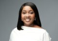 Pennina 1 - The Brief | Namibia's Leading Business & Financial News Portrait of a smiling woman with straight black hair, wearing a white off-the-shoulder top and pearl earrings, against a gray background.