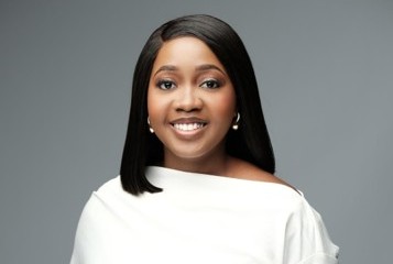 Pennina 1 - The Brief | Namibia's Leading Business & Financial News Portrait of a smiling woman with straight black hair, wearing a white off-the-shoulder top and pearl earrings, against a gray background.