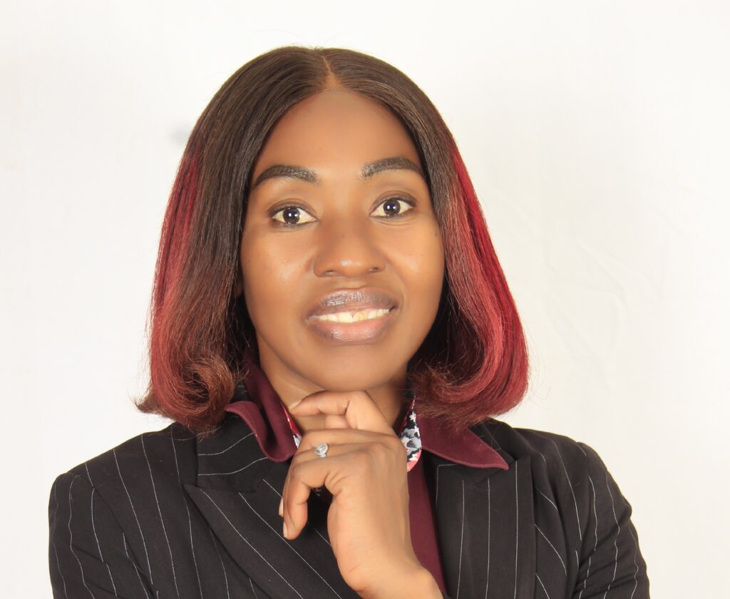 DR HAMBELENI - The Brief | Namibia's Leading Business & Financial News Portrait of a smiling woman with dark hair and red tips, hands clasped under chin, wearing a pinstripe blazer and burgundy scarf against a light backdrop.