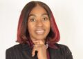 DR HAMBELENI - The Brief | Namibia's Leading Business & Financial News Portrait of a smiling woman with dark hair and red tips, hands clasped under chin, wearing a pinstripe blazer and burgundy scarf against a light backdrop.