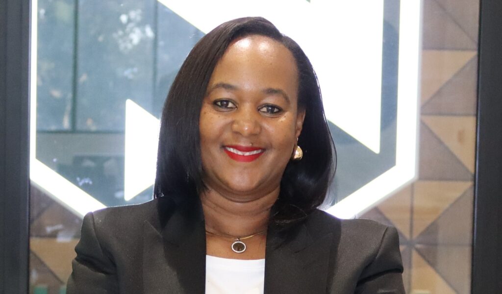 Eunice Tjituka - The Brief | Namibia's Leading Business & Financial News Smiling Black woman in a black blazer with white top, wearing red lipstick and gold earrings, indoors near geometric glass background.