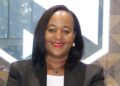 Eunice Tjituka - The Brief | Namibia's Leading Business & Financial News Smiling Black woman in a black blazer with white top, wearing red lipstick and gold earrings, indoors near geometric glass background.