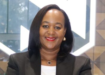 Smiling Black woman in a black blazer with white top, wearing red lipstick and gold earrings, indoors near geometric glass background.