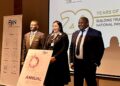 Three professionals in business attire stand on a stage at a PAN Namibia event, beside an 'Annual Report 2025' sign.