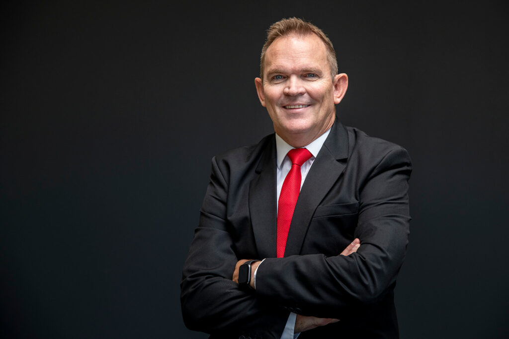 Leon Koch - The Brief | Namibia's Leading Business & Financial News Professional portrait of a man in a black suit and red tie with arms crossed against a dark background.