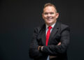Professional portrait of a man in a black suit and red tie with arms crossed against a dark background.