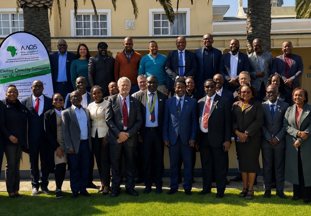 Large group of professionals in business attire posing outdoors in front of a yellow building, with a banner on the left about Uniting Quantity Surveyors on the African Continent