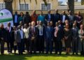WhatsApp Image 2026-07-28 at 50717 PM - The Brief | Namibia's Leading Business & Financial News Large group of professionals in business attire posing outdoors in front of a yellow building, with a banner on the left about Uniting Quantity Surveyors on the African Continent