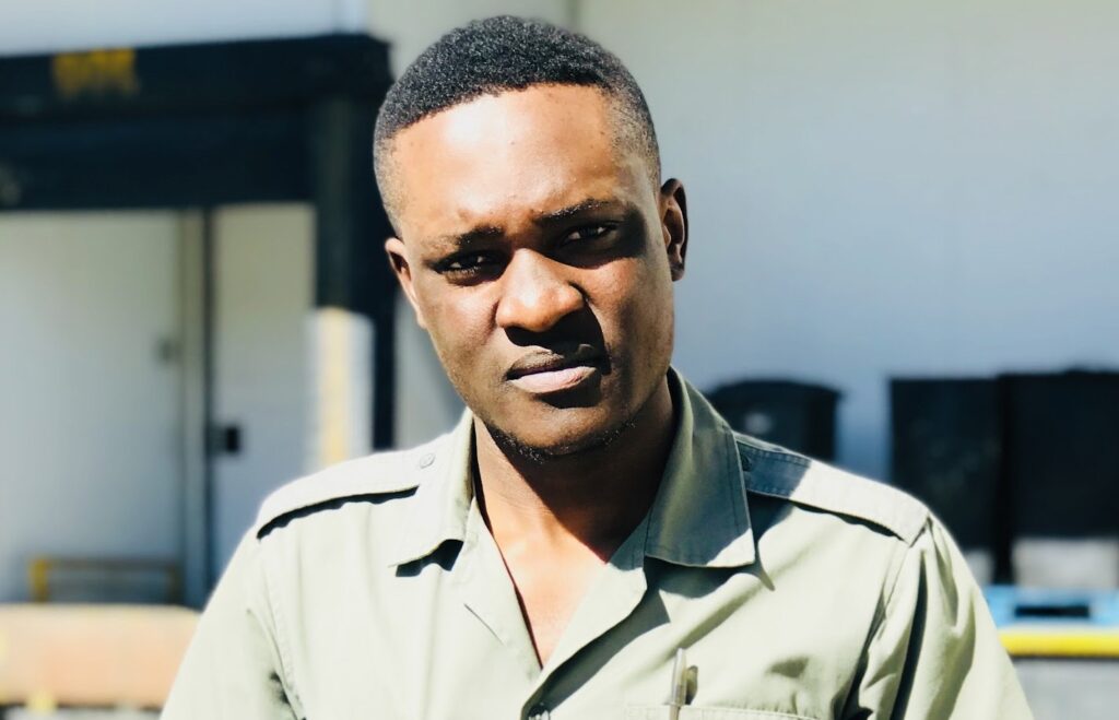 Portrait of a man with short hair in a khaki shirt, squinting slightly in bright sunlight outdoors.