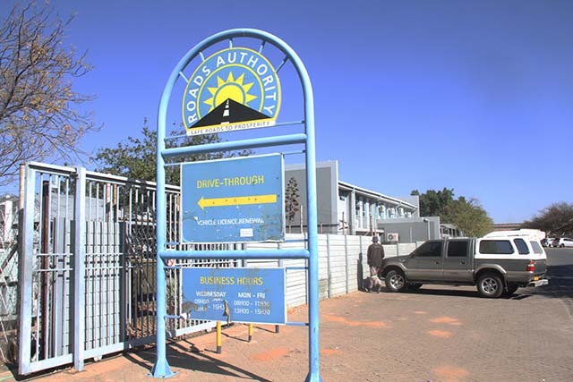 ROADS AUTHORITY - The Brief | Namibia's Leading Business & Financial News Blue metal arch sign for a Road Authority drive-through vehicle license renewal, with a parked SUV and a person in the background near the building.