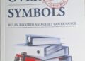 Book cover for 'Systems Over Symbols' with a red 'CONFIDENTIAL' stamp over the title and a stack of binders, folders, and coins on a surface beneath one another, against a light background.