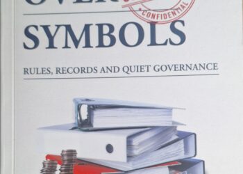 Book cover for 'Systems Over Symbols' with a red 'CONFIDENTIAL' stamp over the title and a stack of binders, folders, and coins on a surface beneath one another, against a light background.
