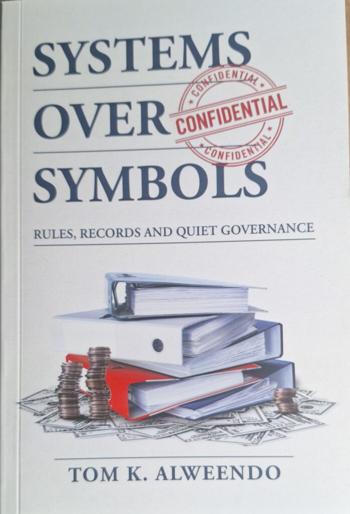 Book cover for 'Systems Over Symbols' with a red 'CONFIDENTIAL' stamp over the title and a stack of binders, folders, and coins on a surface beneath one another, against a light background.