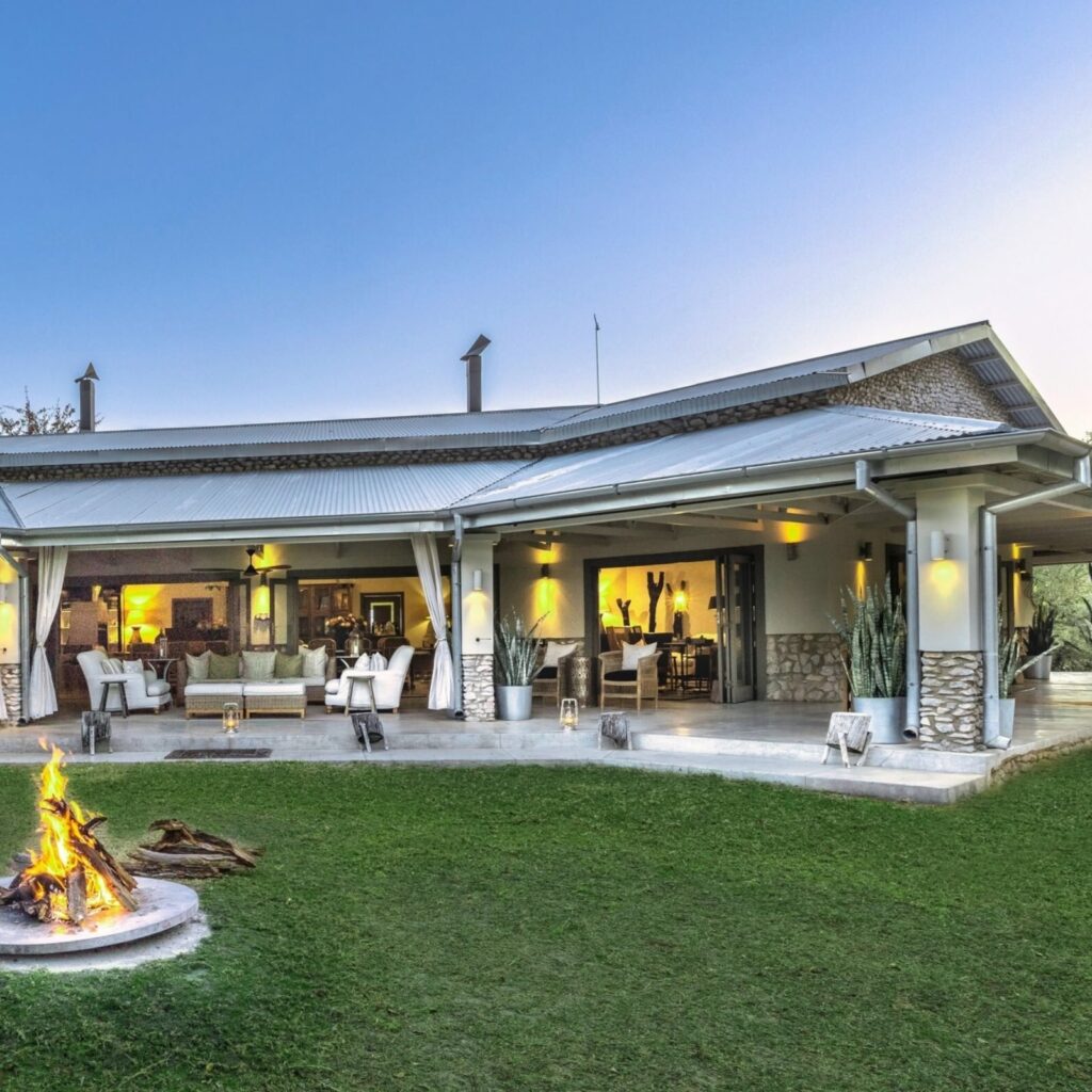 Outdoor patio with white seating, a fire pit on the lawn, and a modern house with large glass doors open to an interior seating area at dusk.