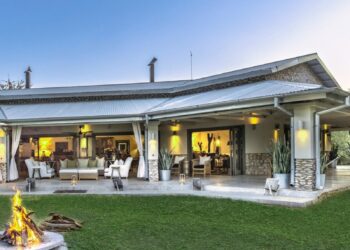 Outdoor patio with white seating, a fire pit on the lawn, and a modern house with large glass doors open to an interior seating area at dusk.