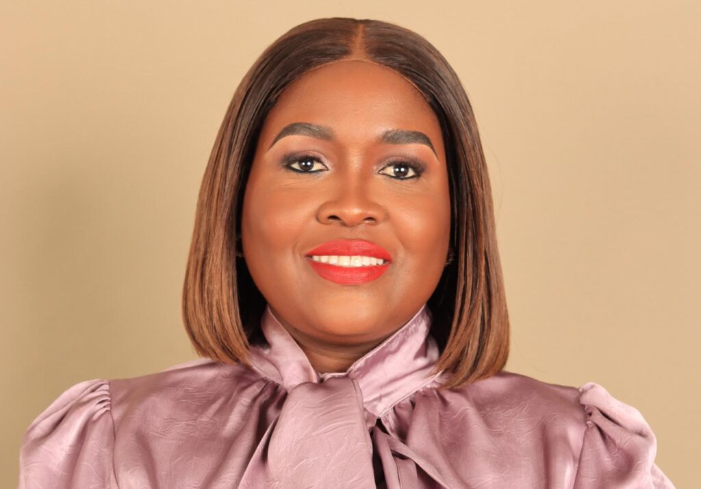 Smiling woman with a shoulder-length brown bob, wearing a pink satin blouse against a beige background.
