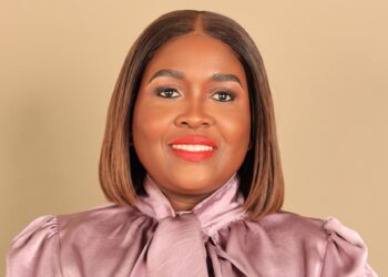 Smiling woman with a shoulder-length brown bob, wearing a pink satin blouse against a beige background.