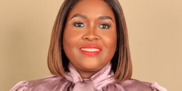 Smiling woman with a shoulder-length brown bob, wearing a pink satin blouse against a beige background.