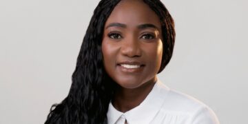 Portrait of a smiling Black woman with long braided hair in a white collared shirt against a light gray background.