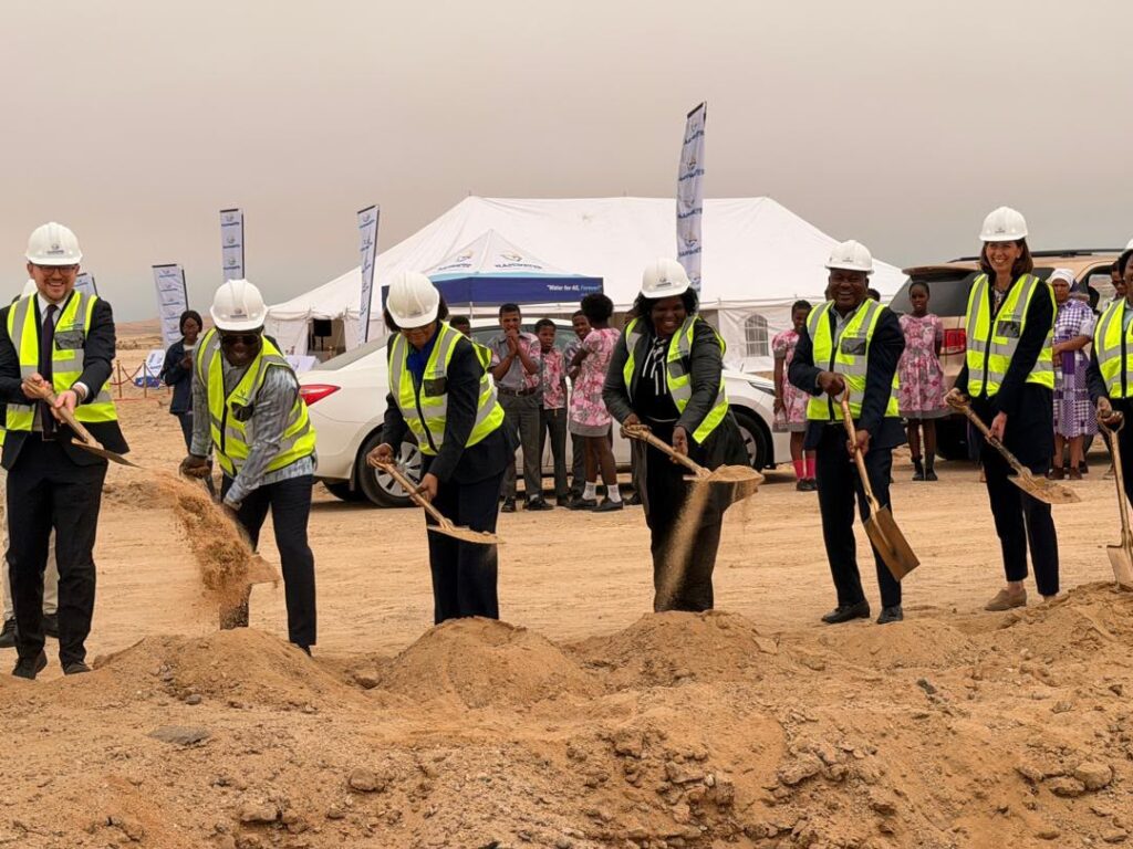 WhatsApp Image 2026-07-30 at 102731 AM - The Brief | Namibia's Leading Business & Financial News Group of professionals in hard hats and high-visibility vests dig with shovels at a desert groundbreaking ceremony, with a white tent and onlookers in the background.