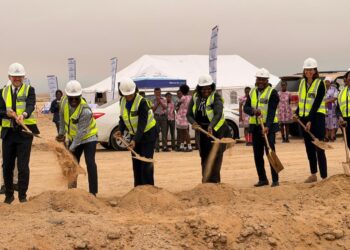 Group of professionals in hard hats and high-visibility vests dig with shovels at a desert groundbreaking ceremony, with a white tent and onlookers in the background.