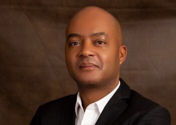 Professional headshot of a man in a black blazer and white shirt against a brown backdrop.