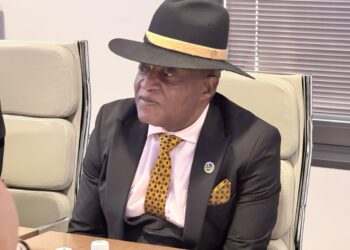 Man in a black suit with a yellow polka-dot tie and fedora, seated at a conference table and looking to the side.