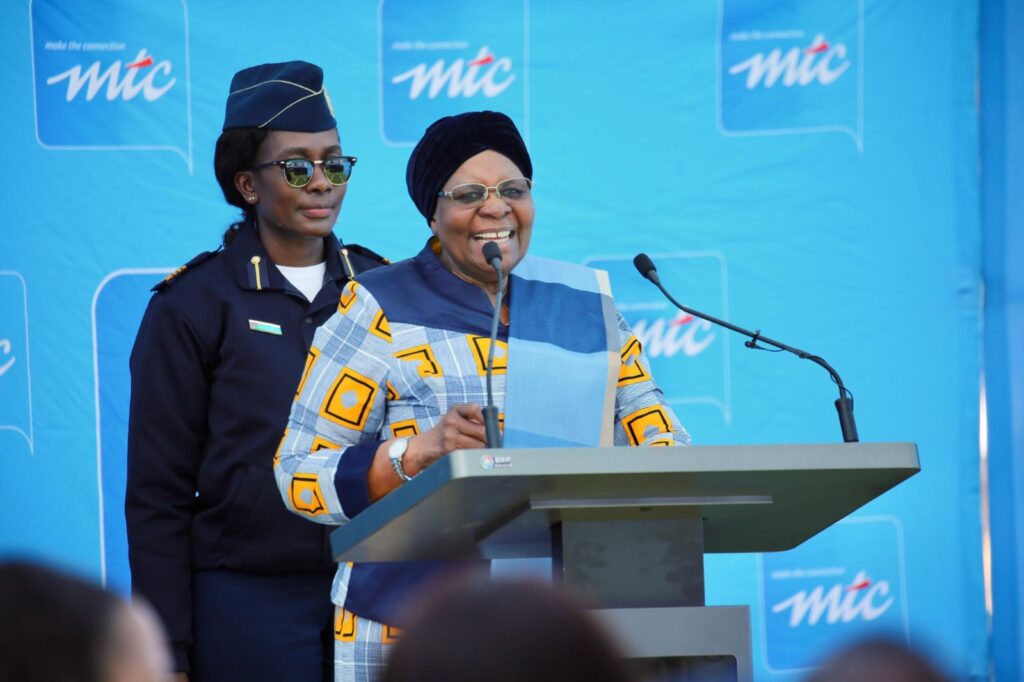 Netumbo 20 - The Brief | Namibia's Leading Business & Financial News Smiling woman in a blue patterned outfit speaks at a podium, with a uniformed aide behind her and a blue backdrop displaying logos behind them.