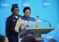 Smiling woman in a blue patterned outfit speaks at a podium, with a uniformed aide behind her and a blue backdrop displaying logos behind them.