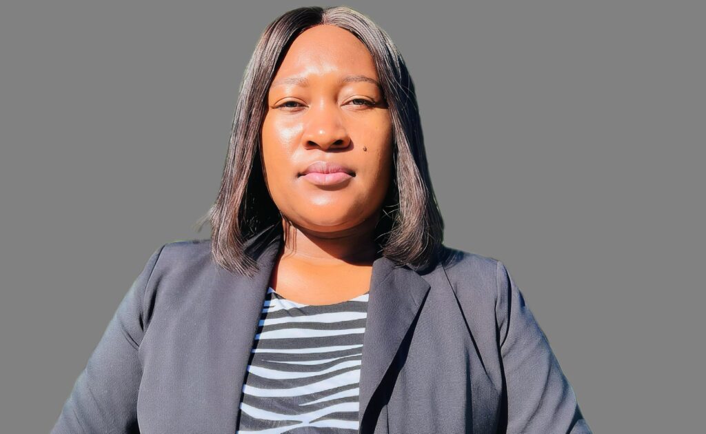 FRIEDA NANGOLO 2 - The Brief | Namibia's Leading Business & Financial News Close-up portrait of a woman wearing a gray blazer over a black-and-white striped top, facing the camera against a gray background.