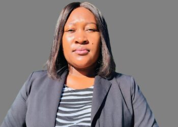 Close-up portrait of a woman wearing a gray blazer over a black-and-white striped top, facing the camera against a gray background.