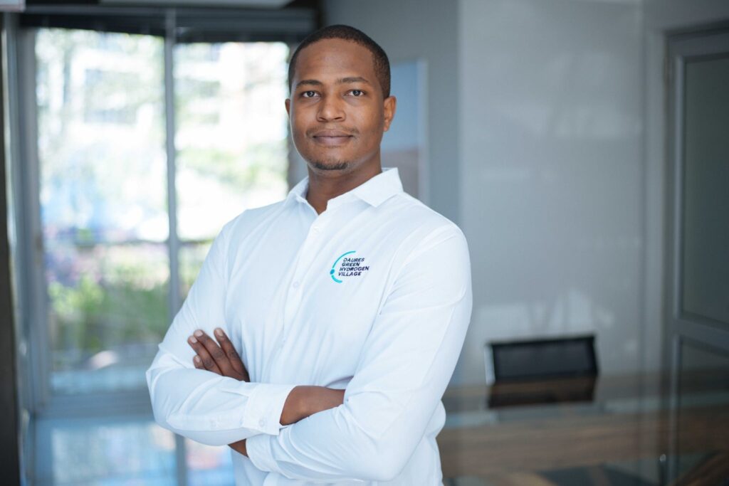 Professional man in a white shirt with a teal logo stands with arms crossed in a bright, modern office setting (logo reads 'DAURES GREEN HYDROGEN VILLAGE').