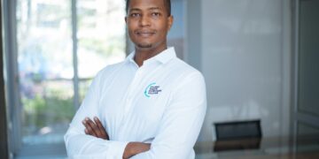 Professional man in a white shirt with a teal logo stands with arms crossed in a bright, modern office setting (logo reads 'DAURES GREEN HYDROGEN VILLAGE').