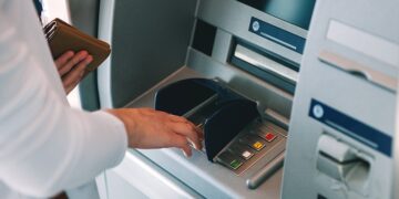 Person using an ATM, inserting a card while holding a wallet nearby in a bank lobby